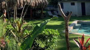 A tropical garden with a thatched-roof structure, a pool, and a white building with black shutters. - Tiki Ora Lodge - HURA  (Bora Bora)