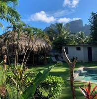 Un jardín tropical con una estructura de techo de paja, piscina y un edificio blanco con contraventanas negras.