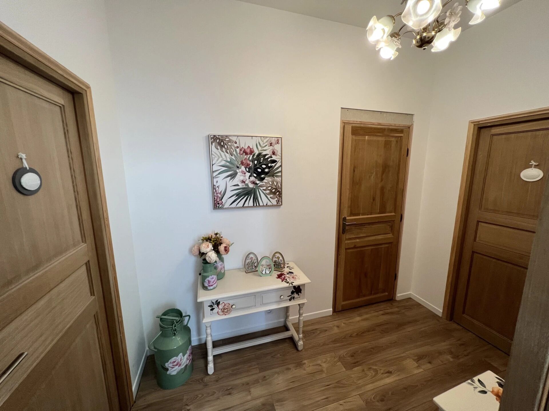Un couloir avec des portes en bois, une petite table ornée d’objets décoratifs et un tableau floral accroché au mur.
