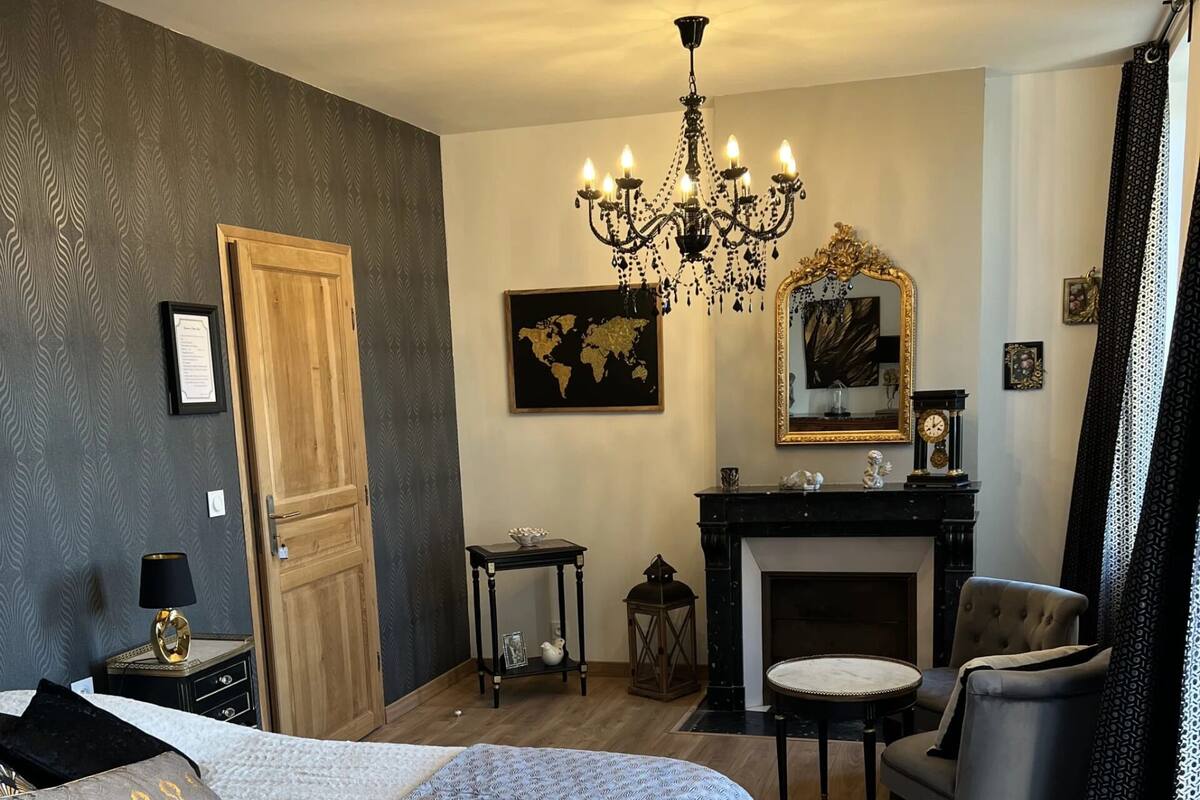 A bedroom with a large bed, a sitting area with a chair and a small table, a fireplace, and a chandelier.
