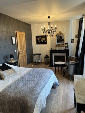 A bedroom with a large bed, a sitting area with a chair and a small table, a fireplace, and a chandelier. - Château Ségot (Neuilly-le-Réal)
