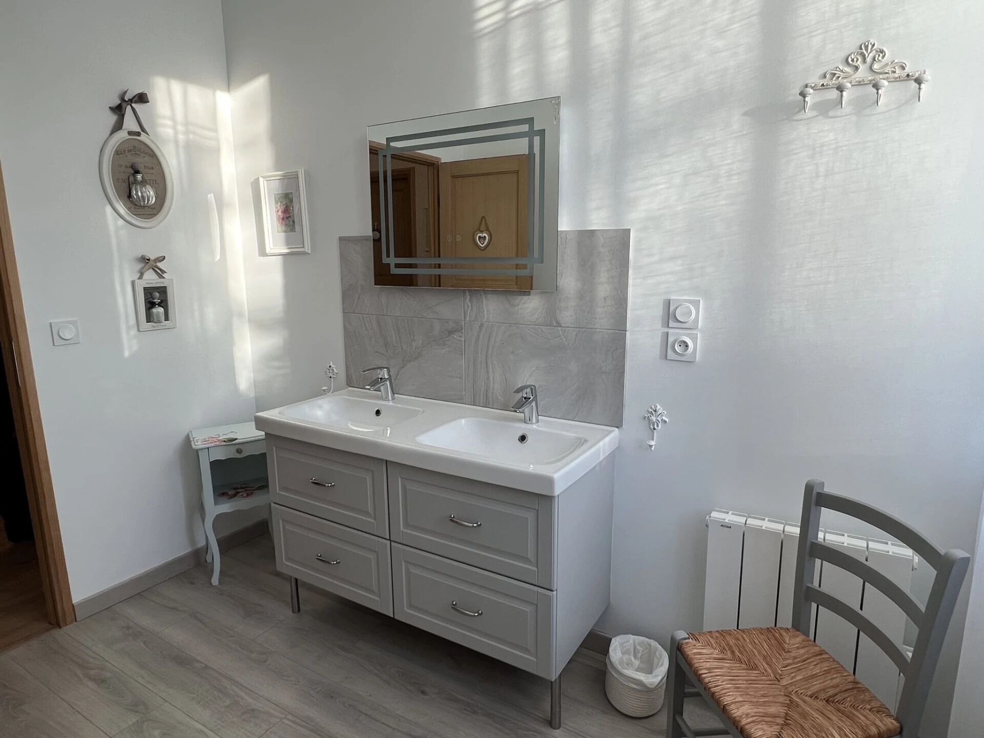 A bathroom with a double sink vanity, a mirror, a small bedside table, and a chair.