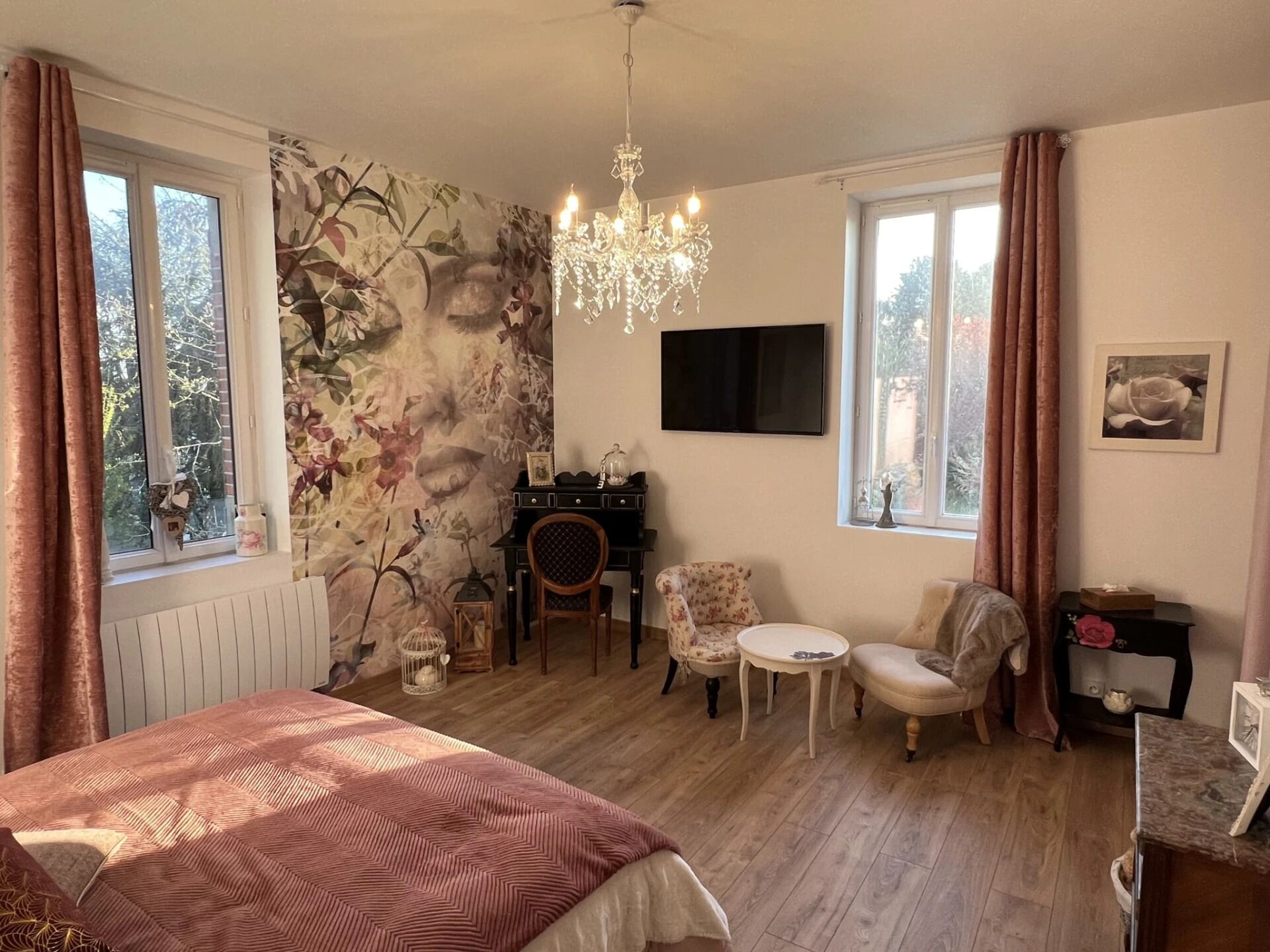 A cozy bedroom with a large bed, a small table, two chairs, a chandelier, and a wall-mounted TV.
