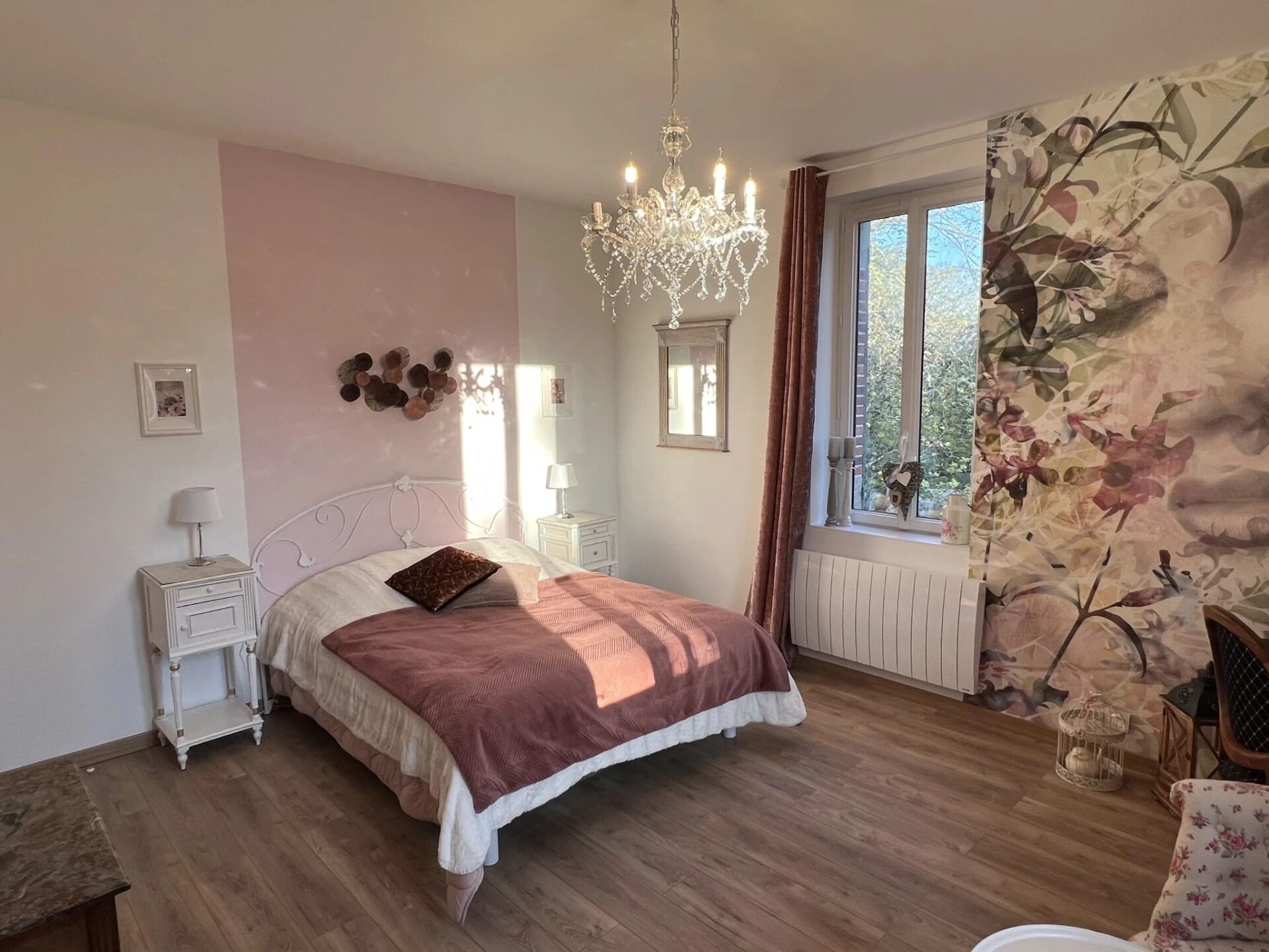 A bedroom with a bed, bedside tables, a chandelier, a window with curtains, and a floral wallpaper.