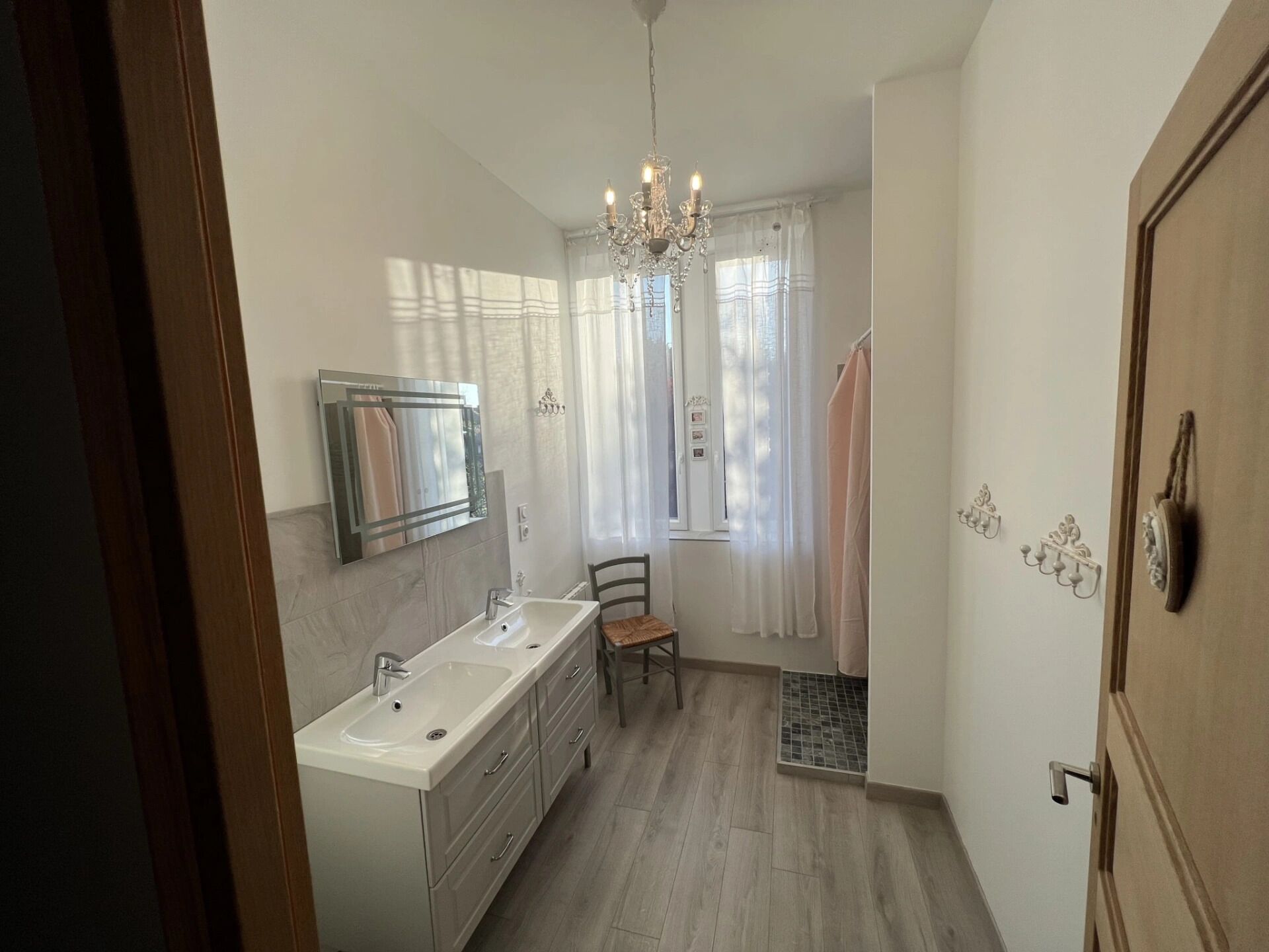 A modern bathroom with a double sink, a mirror, a chandelier, and a shower area.