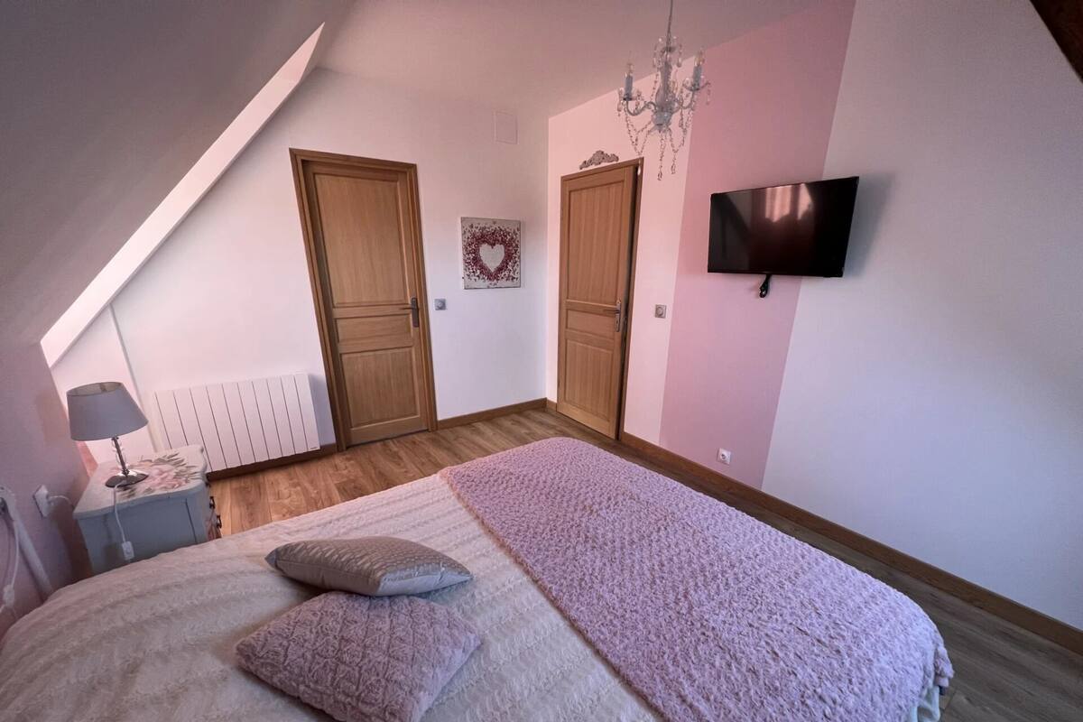 A bedroom with a sloped ceiling, a bed with a pink blanket and pillows, a TV on the wall, and two wooden doors.