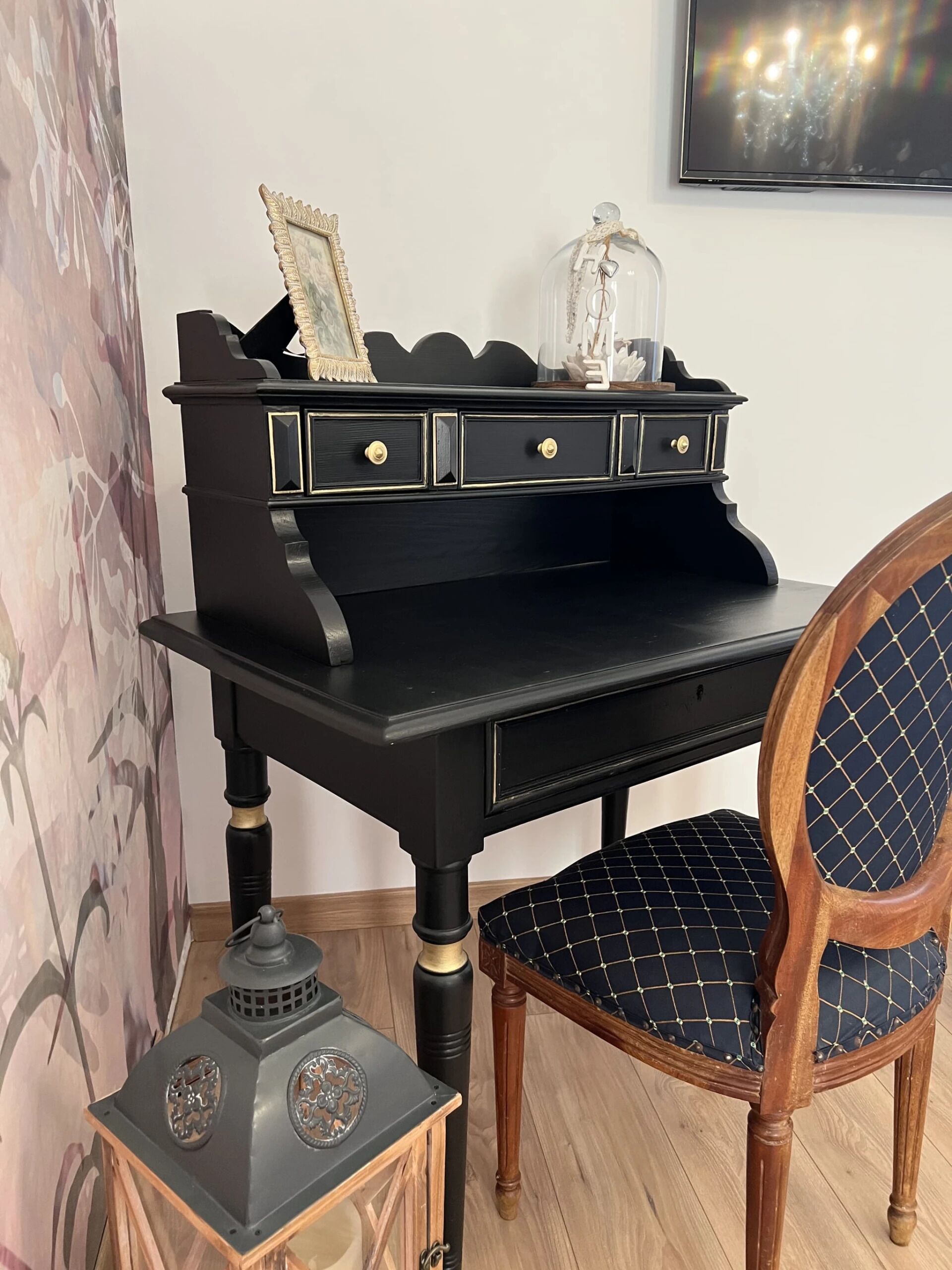 A black desk with three drawers, a lamp, and a chair with a patterned cushion.