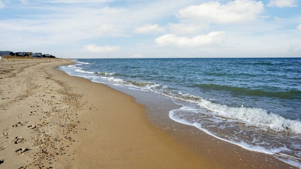 Une plage de sable fin aux vagues douces, un ciel dégagé et une côte lointaine parsemée de bâtiments.