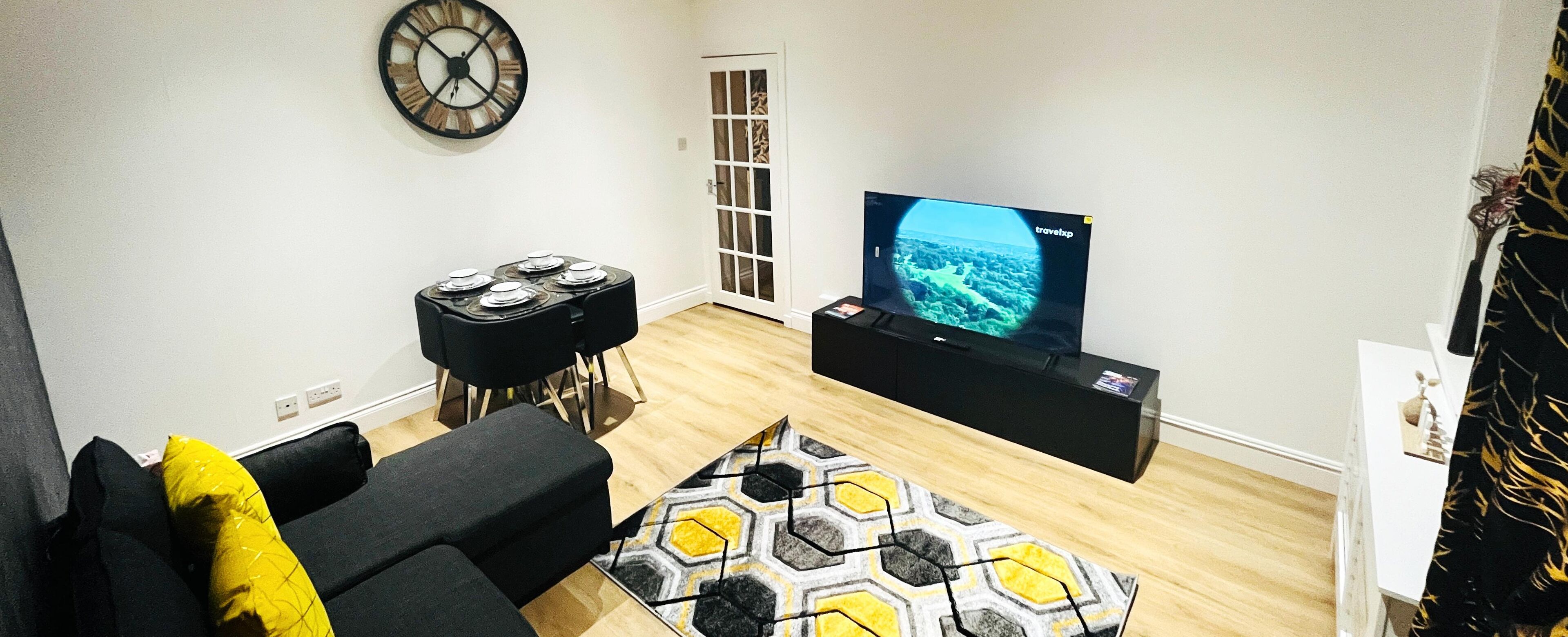 A living room with a black sofa, a yellow pillow, a black coffee table, a black TV stand with a TV displaying a travel channel, and a patterned rug.