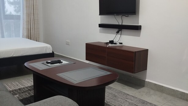 A modern hotel room with a bed, a flat-screen TV mounted on the wall, a wooden cabinet, a round coffee table, and a gray sofa.