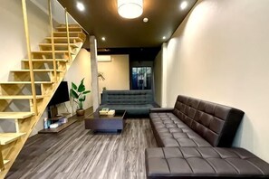 A modern living room with a wooden staircase, a sectional sofa, a coffee table, and a potted plant. - Tachibana (Osaka)