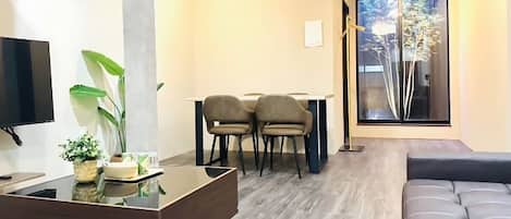 A modern living room with a flat-screen TV, a glass coffee table, a sofa, and a dining area with chairs.
