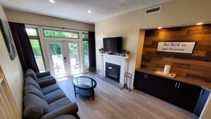 Ein modernes Wohnzimmer mit Sofa, Glastisch, Kamin, Fernseher und Küchenbereich. An der Wand befindet sich ein Schild.
