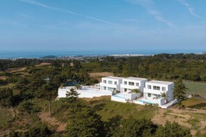 Una villa blanca moderna con piscina, rodeada de vegetación y con vistas a la costa.