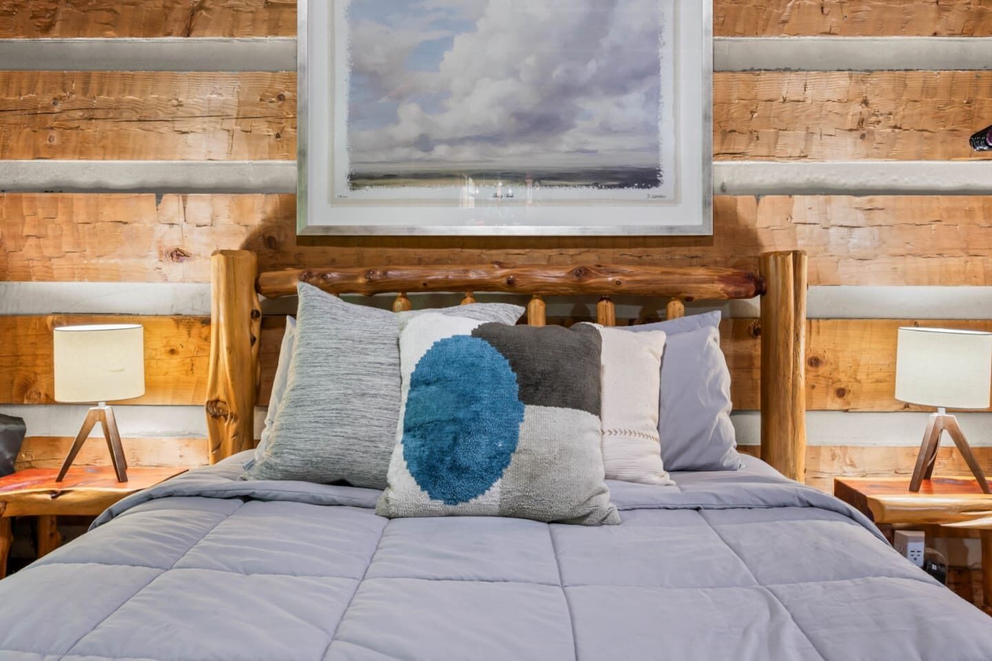 Una cama con colcha gris, cabecera de madera y un cuadro enmarco con un paisaje.