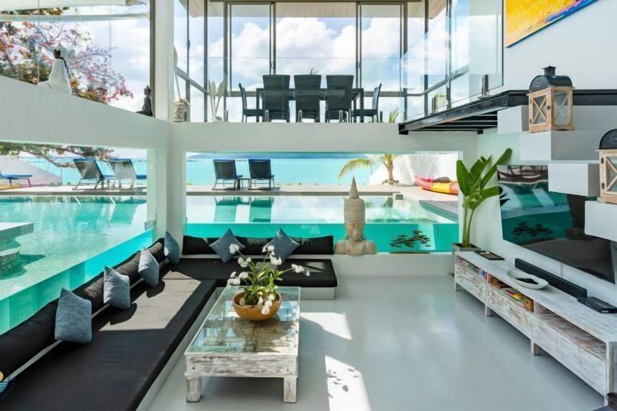 A modern living room with a glass coffee table, a sofa with cushions, a TV stand, and a view of the pool and sky.