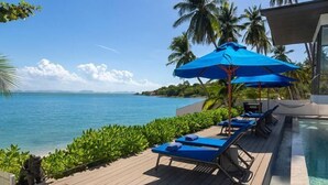 A poolside area with lounge chairs and a blue umbrella, overlooking a clear blue sea with palm trees and a distant shoreline. - Bella Samui Beach Villa (Koh Samui)