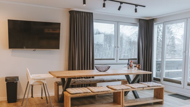A modern living room with a wooden dining table, benches, a TV mounted on the wall, and large windows with curtains. - Vacation apartment Schneeglück for 10 people with large terrace (Winterberg)