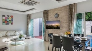 Een moderne woonkamer met een bank, salontafel, eettafel met stoelen, een aan een bakstenen muur bevestigde televisie en uitzicht op een zwembad.