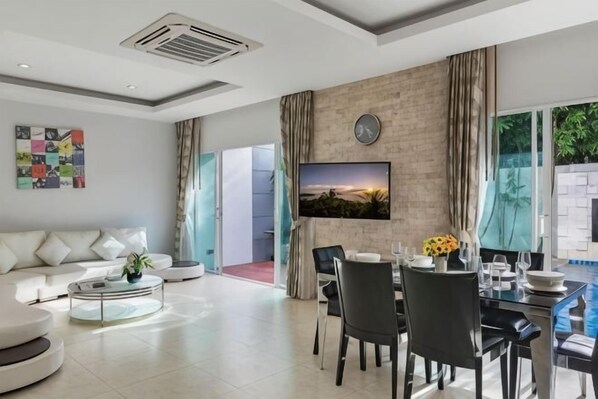 A modern living room with a sofa, coffee table, dining table with chairs, a TV mounted on a brick wall, and a view of a swimming pool. - Trixie Town House Unit 2 (Phuket)