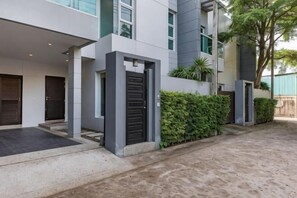 A modern building with a covered entrance, a gated driveway, and a landscaped area with a tree and bushes. - Trixie Town House Unit 2 (Phuket)