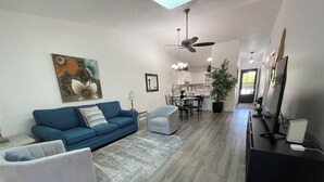 A living room with a blue sofa, grey armchairs, a dining area with a chandelier, and a kitchen in the background.