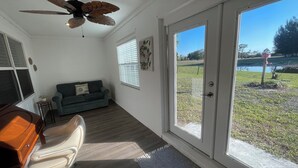 A room with a ceiling fan, a desk with a chair, a couch, and a large sliding glass door leading to an outdoor area with a pond and a birdhouse.