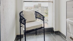 Un balcon avec une chaise en métal noir et un coussin blanc, un mur blanc et une vue sur des immeubles à travers une fenêtre.