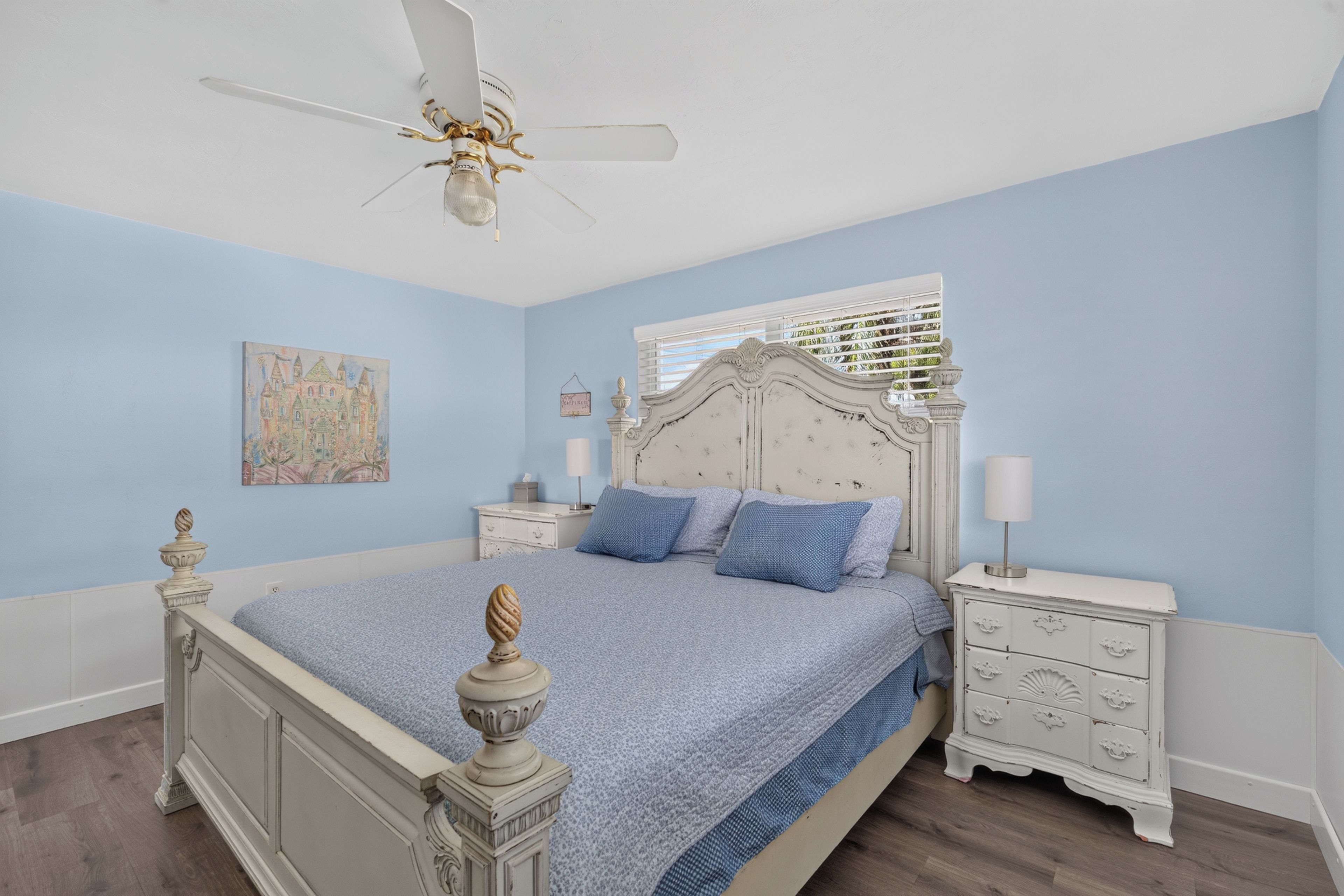 A bedroom with a bed, nightstands, a ceiling fan, a window with blinds, and a wall painting.