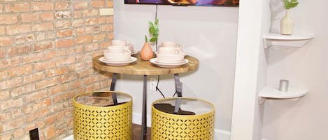 Una habitación con pared de ladrillo, una mesa de madera con tazas y platos, dos contenedores de basura con diseño amarillo y una estantería con una planta.