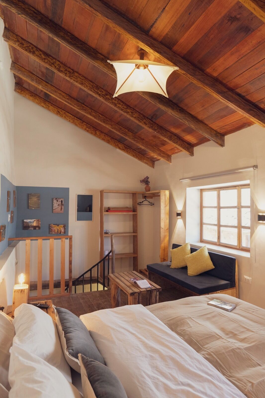 Un dormitorio acogedor con techo de madera, cama con ropa de cama blanca y gris, una mesita y una ventana con vistas.