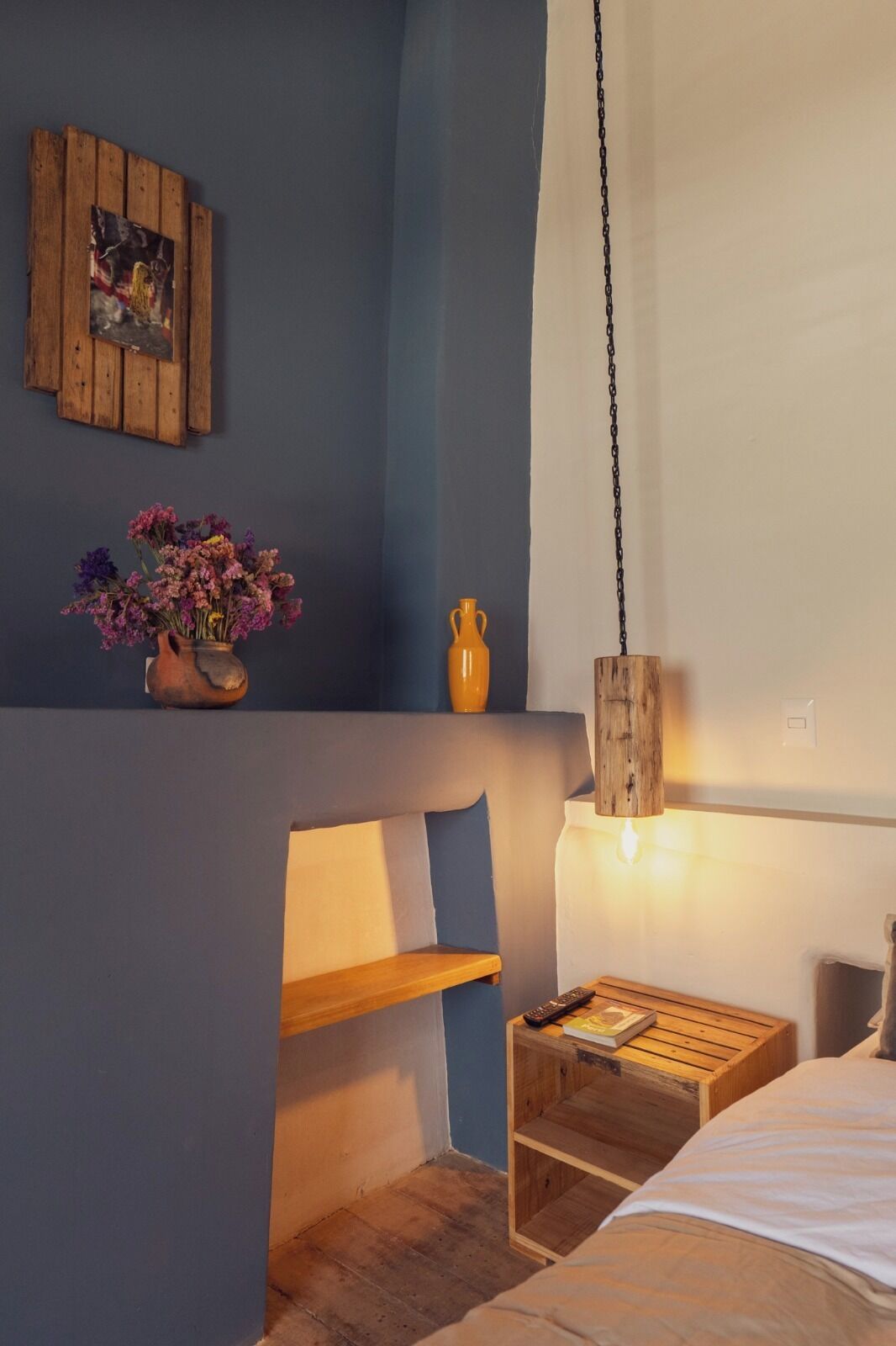 A bedroom with a bed, a bedside table with a lamp, a wooden bench, a vase with flowers, and a painting on the wall.