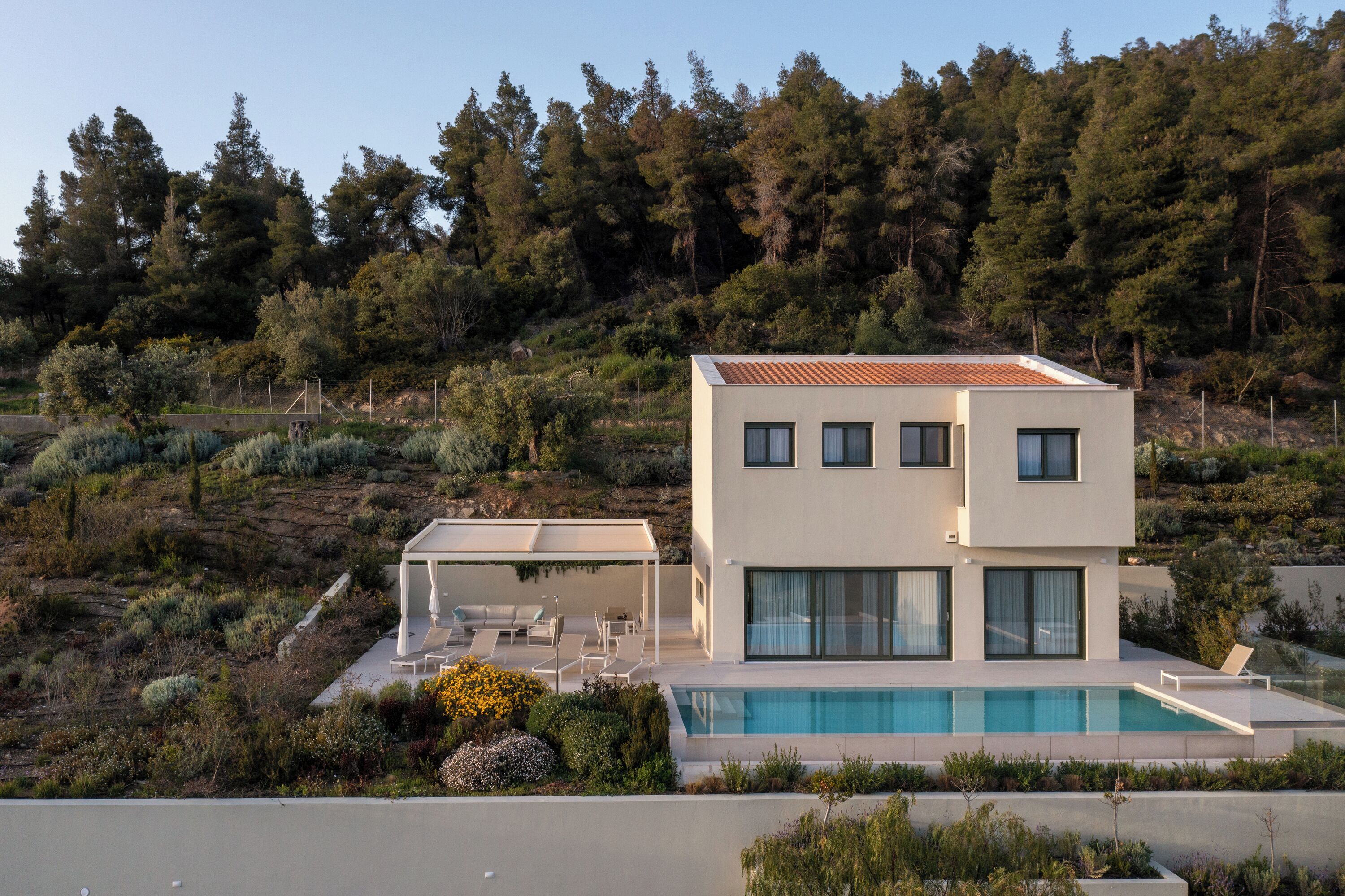 A modern two-story house with a swimming pool, surrounded by a landscaped garden and a forested hillside.