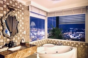 A bathroom with a large bathtub, a sink, a mirror, and a window with a city view. - Empress Dalat Hotel (Dalat)