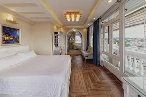 A hotel room with a large bed, wooden flooring, a view through the window, and a door leading to a balcony.