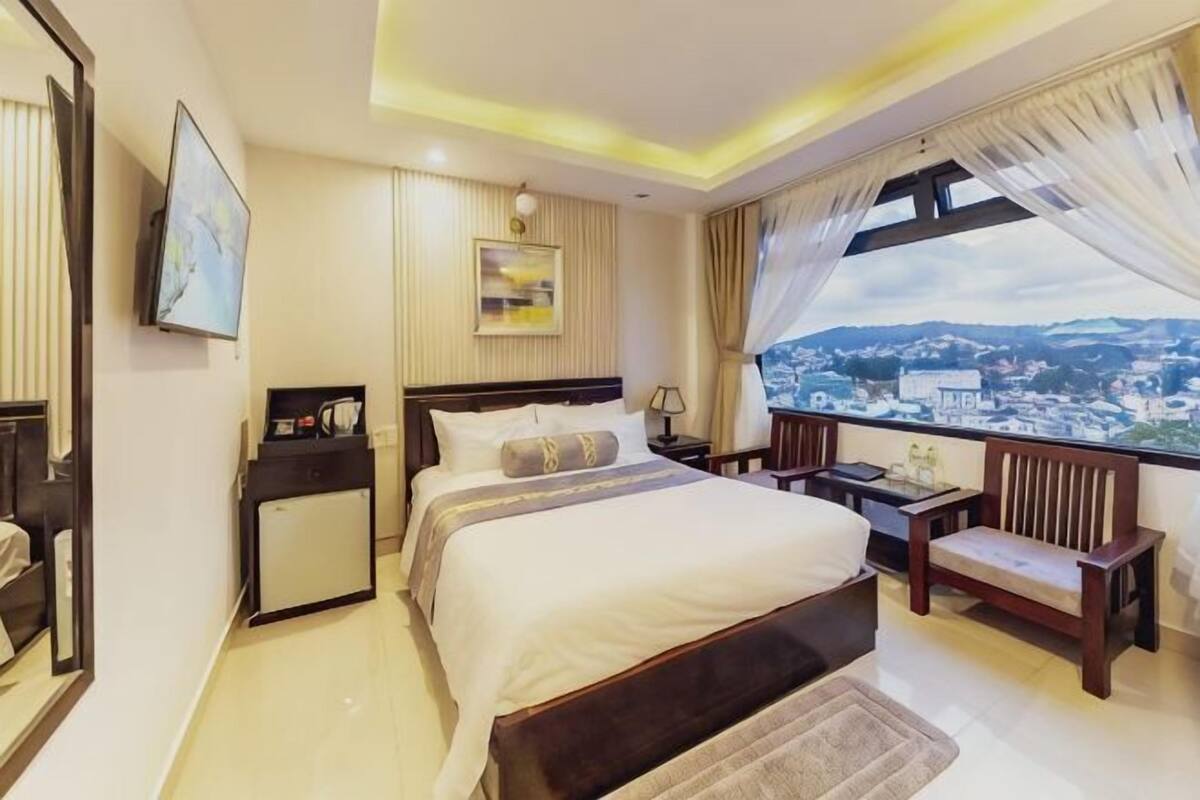 A hotel room with a large bed, bedside tables, a desk, a chair, a view of a cityscape, and a window with curtains.