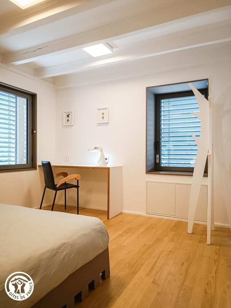 A bedroom with a bed, a desk, and a chair. There are two windows with blinds, a radiator, and a white wall.