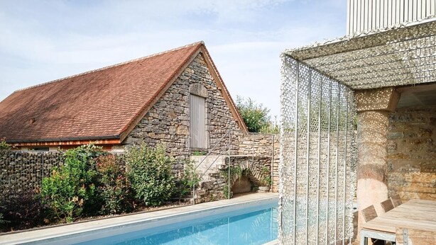 A swimming pool adjacent to a stone building with a tiled roof, surrounded by greenery and a wooden deck with a table and chairs. - Gîtes de France® - Paty (CAJARC)