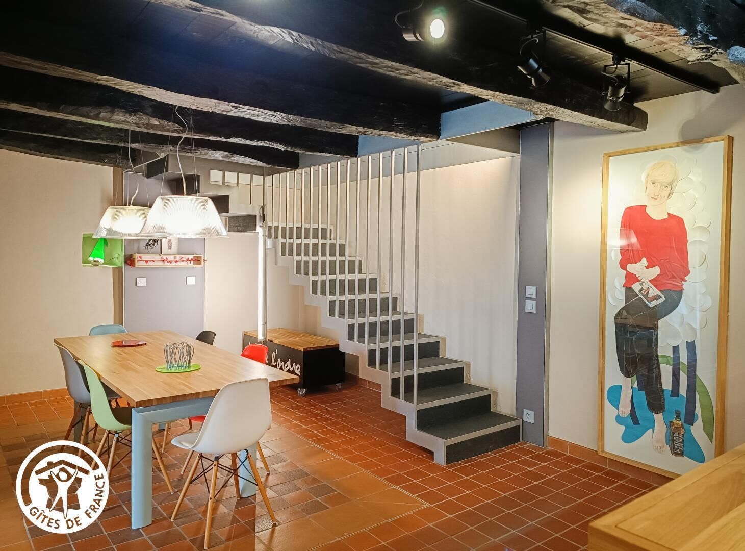 Un intérieur moderne avec une table à manger en bois, des chaises, un escalier et une œuvre d’art accrochée au mur.