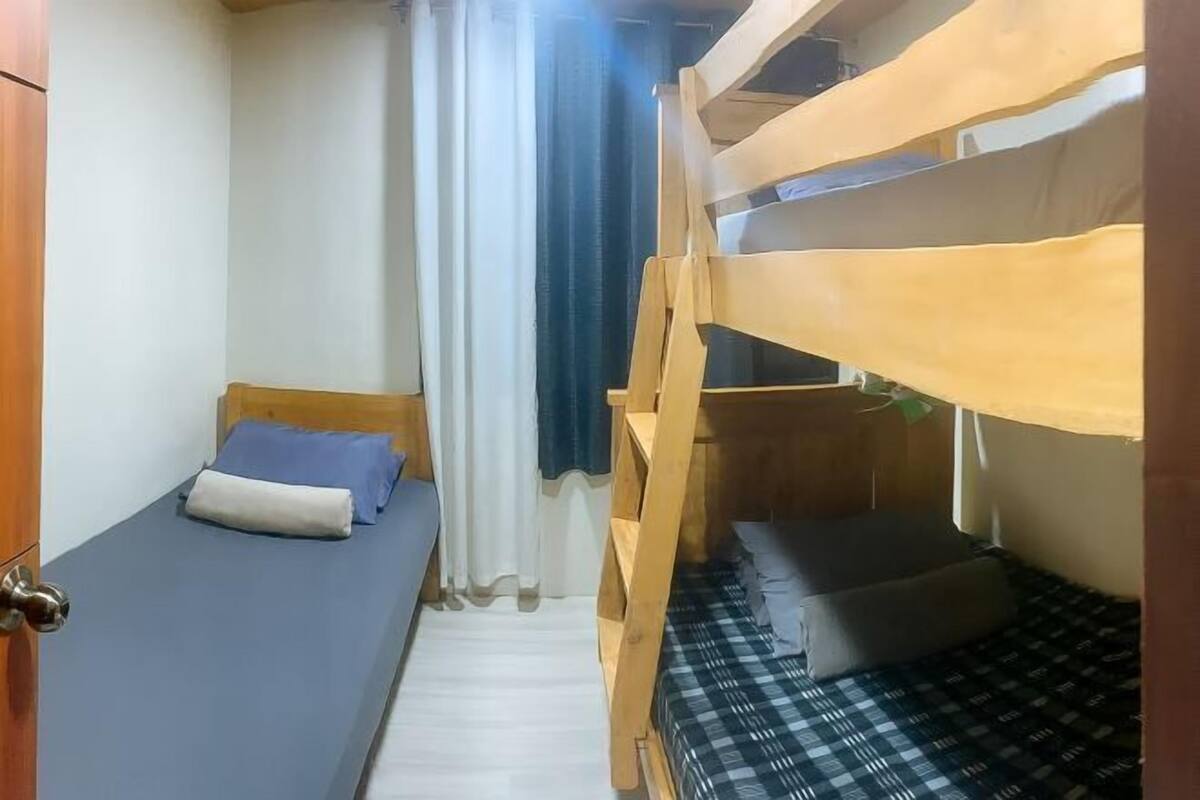 A small, compact room with bunk beds, a single bed, a wooden door, and a curtain.
