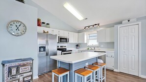 A modern kitchen with white cabinets, a central island with stools, a refrigerator, microwave, and a clock on the wall. - Practically Perfect on West Beach - Just steps away from the Atlantic Ocean (Oak Island)