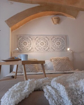 A bedroom with a bed, a wooden table with a cup, a lamp, and a decorative wall panel. - Suite in the Sasso Caveoso (Matera)