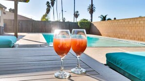 Zwei Gläser rotes Getränk auf einer Holzterrasse an einem Pool, im Hintergrund eine Holzlattenveranda und Palmen.