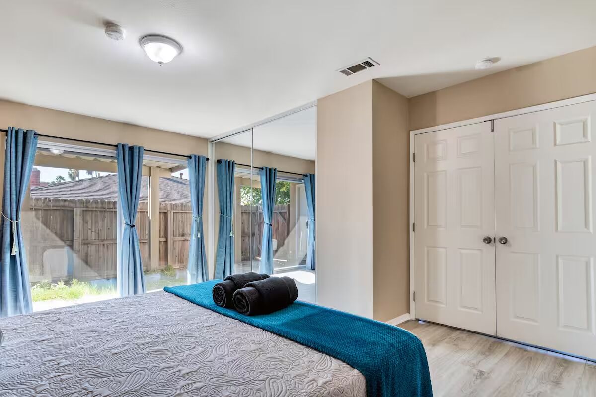 Ein Schlafzimmer mit einem großen Bett, einem Fenster mit Vorhängen, einer Schiebetür und Blick auf den Garten.