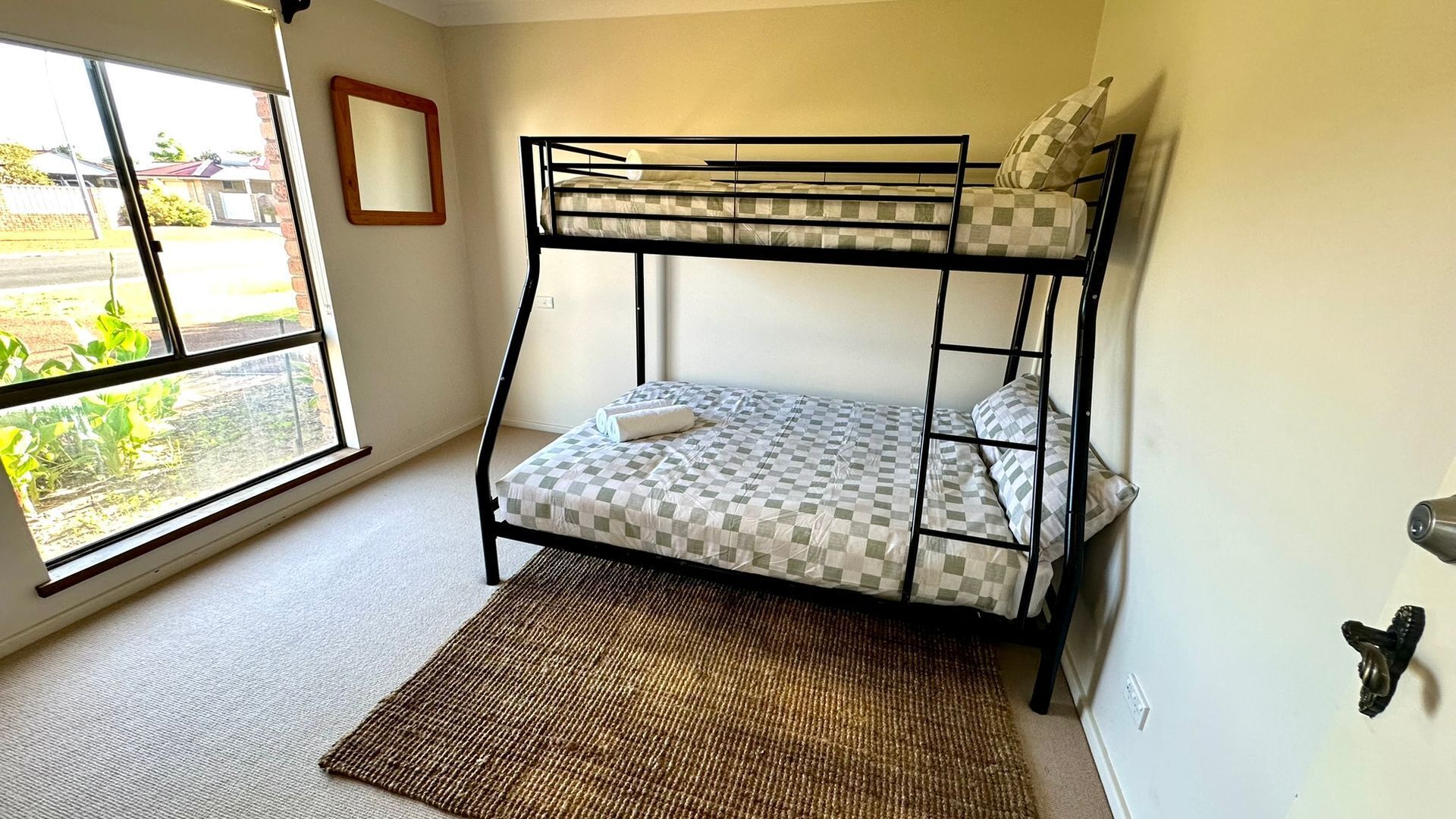 Una cama litera con ropa de cama a cuadros, una ventana con vistas a la vegetación y una alfombra tejida en el suelo.