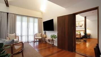 A modern living room with a flat-screen TV, wooden flooring, and a large window with curtains.