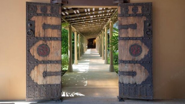 Um corredor com uma porta de madeira na entrada, que dá para uma galeria coberta com colunas.