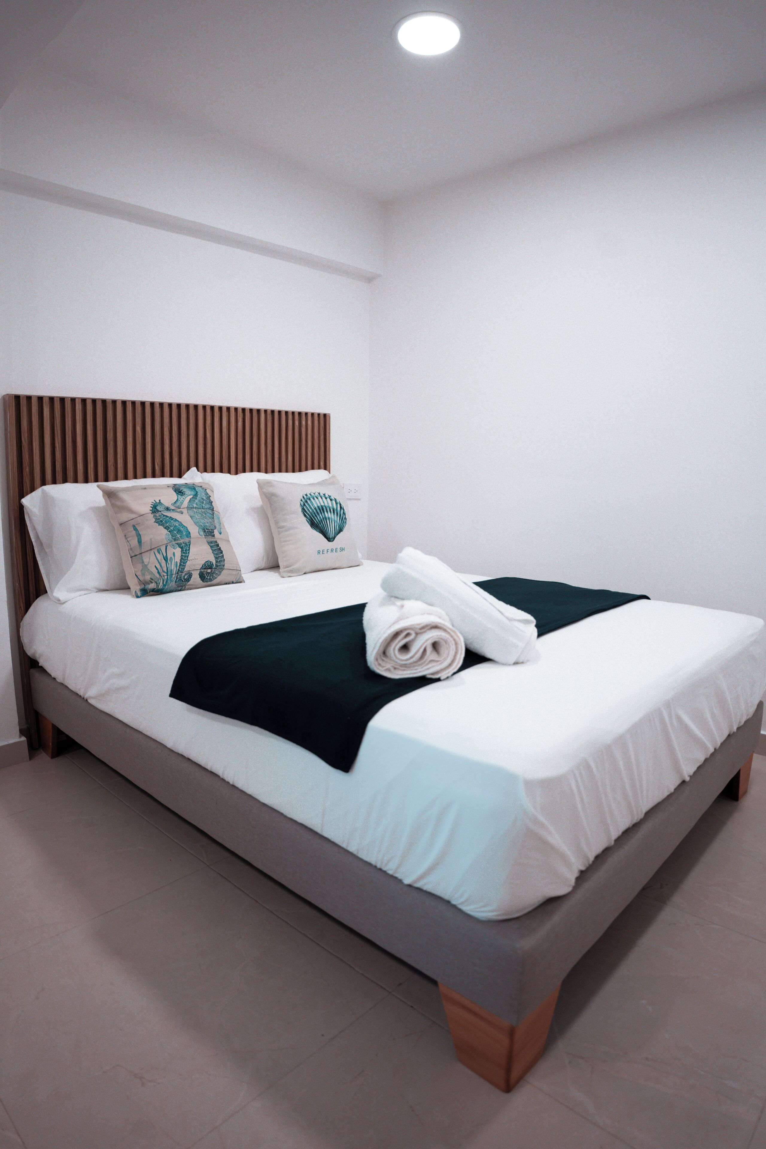 Una cama bien hecha con sábanas blancas, una manta verde oscuro y almohadones decorativos con diseño de caballito de mar y concha.