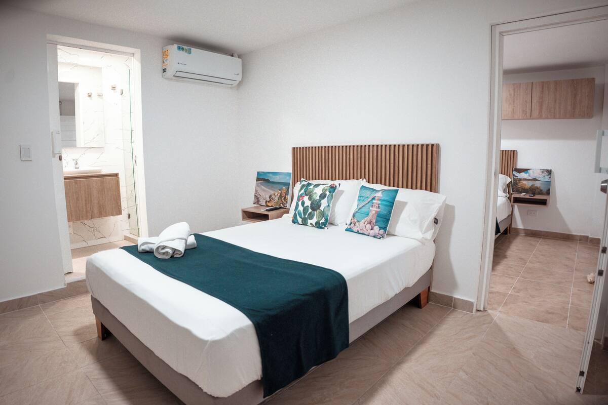 A hotel room with a bed, a nightstand, a wall-mounted air conditioner, and a bathroom visible through an open door.