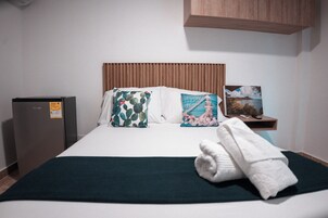 A neatly made bed with a green bedspread, two decorative pillows, a folded white towel, a small wooden shelf with a painting, and a refrigerator to the left.
